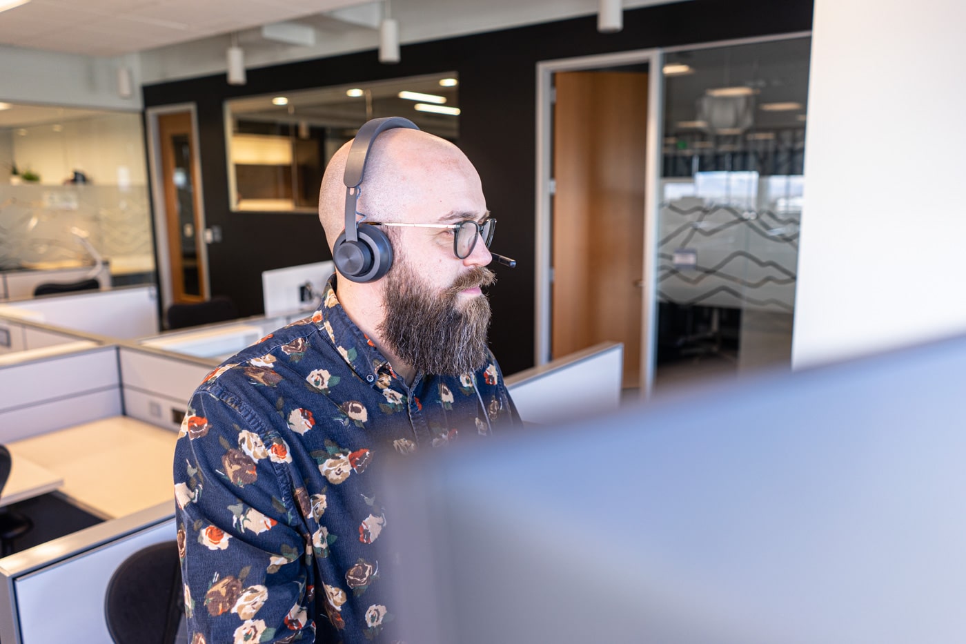white bald man with a beard looking at a computer working with a headset on