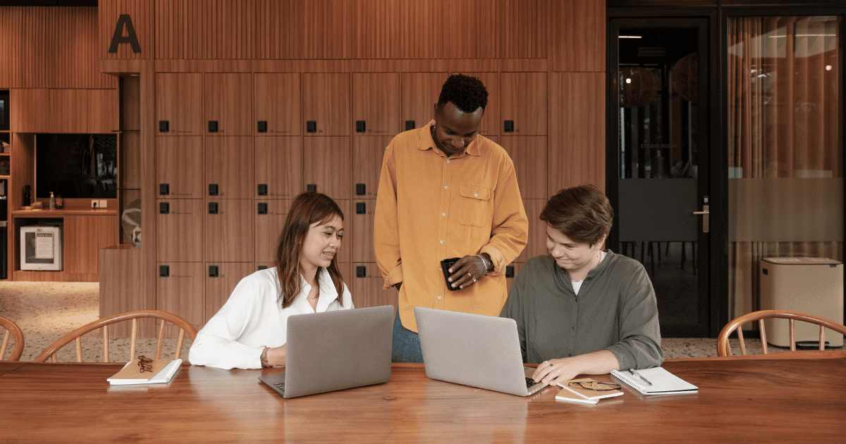 Two people sitting at a table, working on laptops while one guy is standing and looking at them.