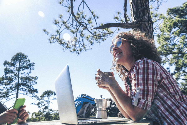 Woman sitting at a table outside with a laptop and a coffee cup in her hand.