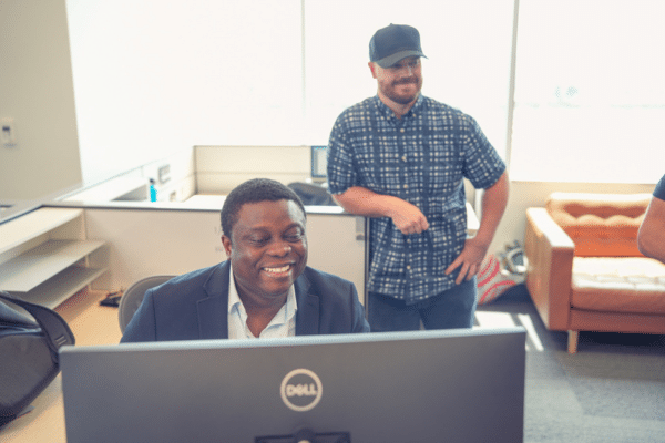 One man sitting at a desk, smiling at a computer. Another man stands behind him.