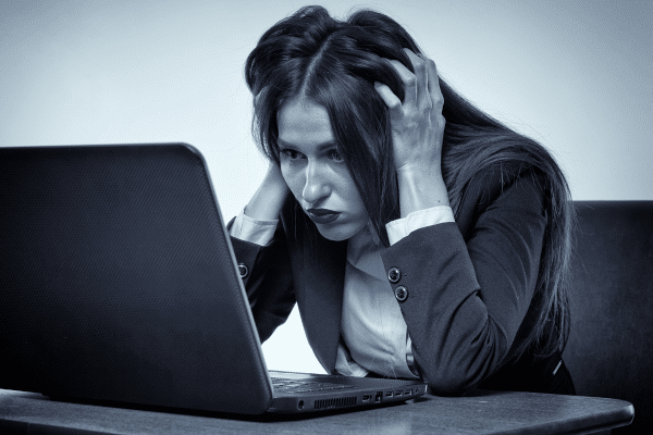 Woman sitting at a desk, hands on her head, looking very frustrated as she glares at her laptop.